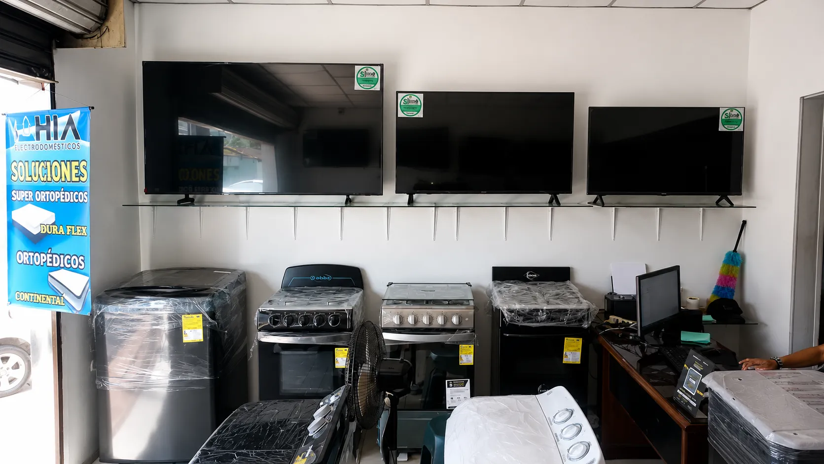 Interior del almacén Bahía: televisores exhibidos en una repisa de vidrio, estufas y lavadoras debajo, junto a un pendón de colchones ortopédicos.